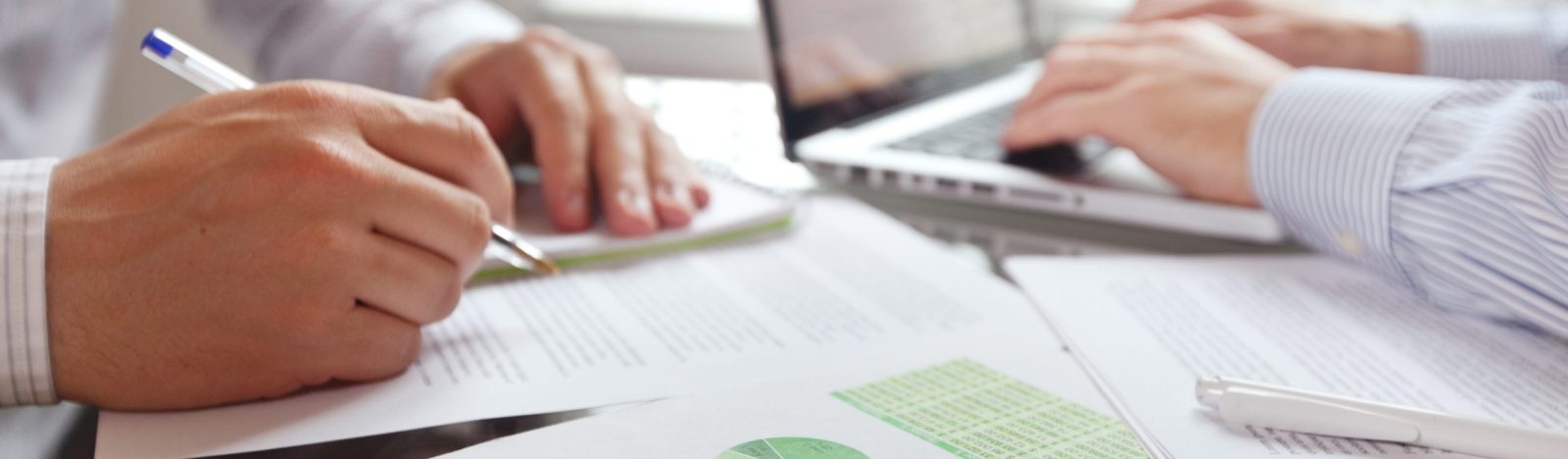Zwei Personen arbeiten an einem Schreibtisch mit Stift und Papier und Laptop. Der Bildfokus ist auf die Hände im Vordergrund gerichtet.